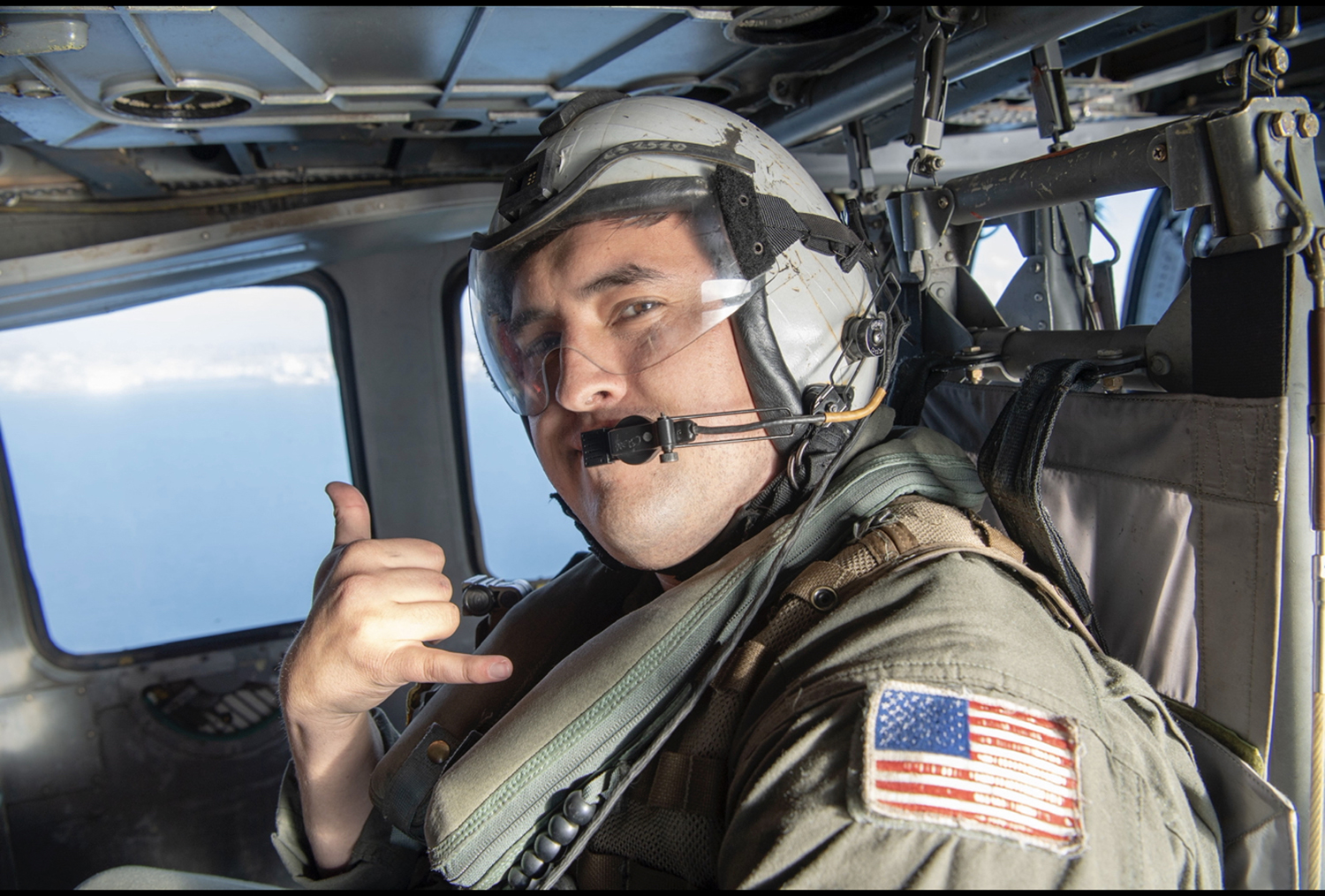 a military student gives the 'hang 10' symbol with his hands (pinky and thumb stuck out) while he flies in a plane in military gear