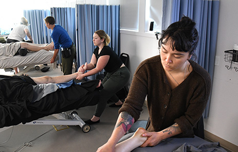 A massage therapy student in an educational setting practices massage on another student's leg while other students practice in the background